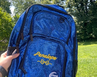 Puede incluir: Una mochila azul brillante con cremalleras y ribetes negros. La mochila tiene el nombre "Penelope" bordado en escritura amarilla con un diseño de lazo. La marca "Chassé" también es visible en la parte delantera.