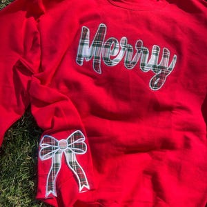Pode incluir: Camisola vermelha com um design "Merry" xadrez e um laço xadrez na manga.