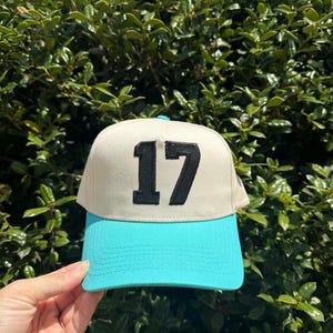 May include: A baseball cap with a cream-colored crown and a turquoise brim. The number "17" is embroidered in black on the front of the cap. The cap is held up against a backdrop of green foliage.