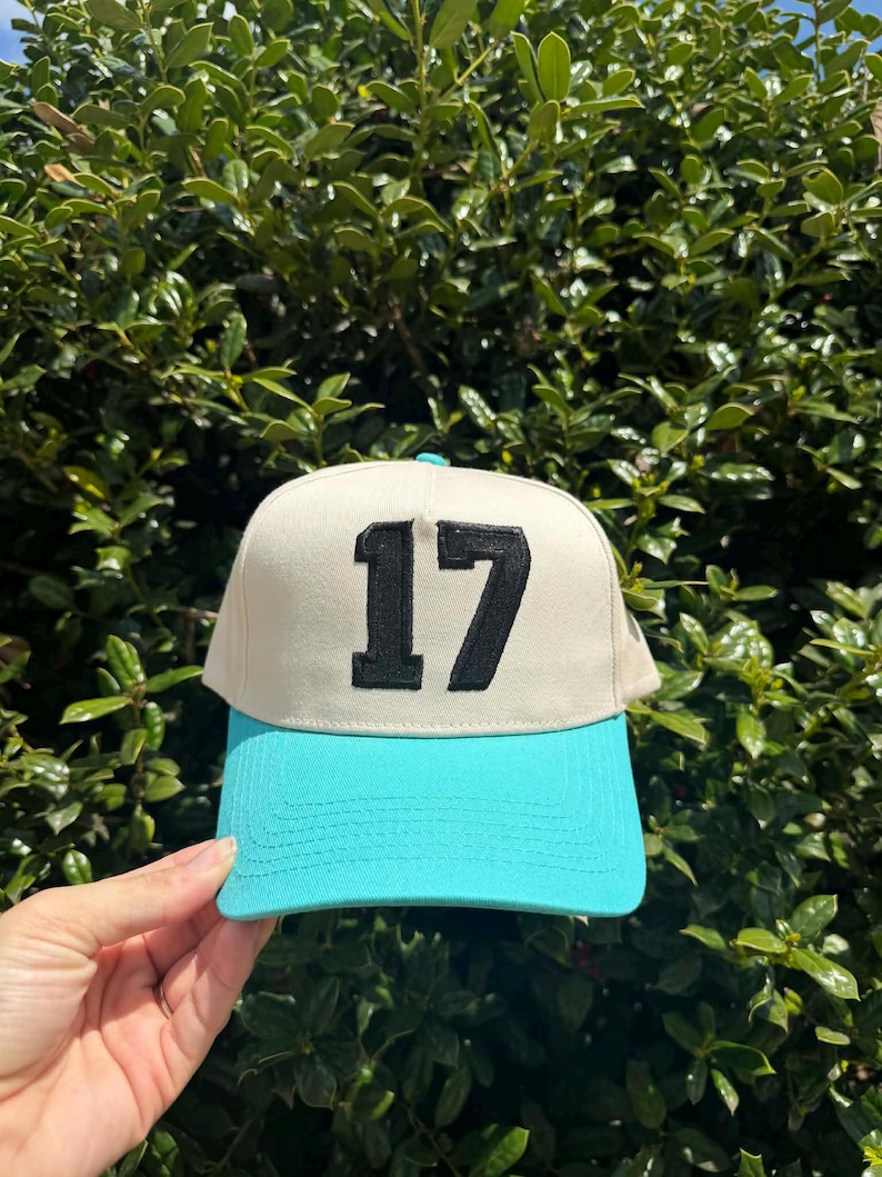 May include: A baseball cap with a cream-colored crown and a turquoise brim. The number "17" is embroidered in black on the front of the cap. The cap is held up against a backdrop of green foliage.