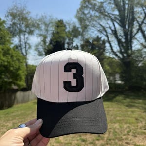 Pu&ograve; includere: Un cappellino da baseball con visiera nera e corona bianca a sottili righe verticali. Il numero "3" &egrave; mostrato in nero sul davanti. Il cappellino &egrave; tenuto contro uno sfondo di alberi verdi e cielo blu.
