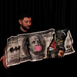 May include: A large, folded artwork resembling a $100 bill, featuring Benjamin Franklin with a bubblegum bubble. The design incorporates Batman imagery and comic book panels, with a black background.