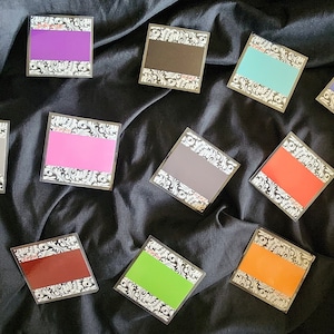 Puede incluir: Un conjunto de 12 tarjetas cuadradas de colores con un borde decorativo. Las tarjetas están dispuestas en semicírculo sobre un fondo de tela negro. Las tarjetas están etiquetadas con colores, incluyendo rojo, naranja, amarillo, verde, azul, morado y gris. Una tarjeta está etiquetada con "Wild".