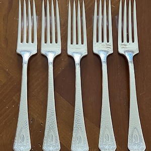 May include: Five silver forks with tines and textured handles. Each fork has a decorative design at the base of the handle. The forks are arranged in a row on a wooden surface. The forks are a classic design.
