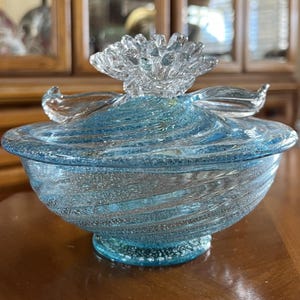 May include: A decorative glass bowl with a lid, featuring a swirling blue and clear design. The bowl has a round base and a clear, flower-shaped finial on top. The glass has a textured, swirled pattern.
