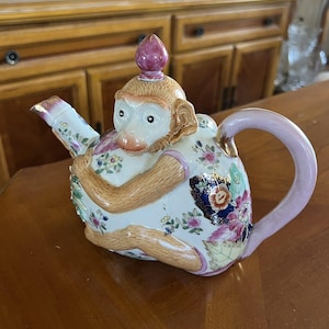 May include: A ceramic teapot shaped like a monkey with a pink and white floral design. The teapot has a pink handle and a gold spout.