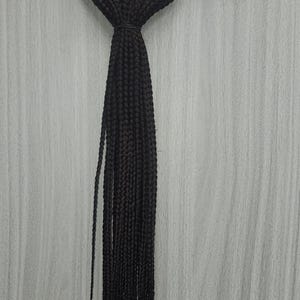 May include: Black braided hair extensions hanging from a wire hanger. The hair is tightly woven into small braids, with a knot at the top and a rubber band securing the braids. The hair extensions are a solid black color.