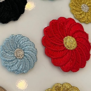 Puede incluir: Surtido de seis parches de flores de tela bordados en varios colores, incluyendo morado, rojo, azul, negro, dorado y azul claro. Cada flor tiene un centro plateado y un centro amarillo.