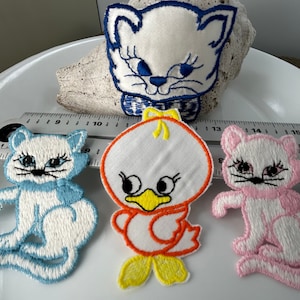 May include: Three embroidered iron-on patches featuring a white cat with a blue scarf, a white duck with orange and yellow accents, and a pink cat with white accents.