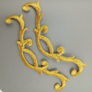 May include: Two gold embroidered fabric embellishments in the shape of stylized floral branches. The embellishments are intricate and detailed, with a delicate, flowing design.