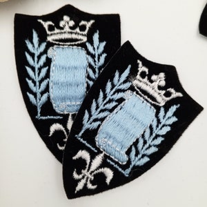 May include: Two black velvet patches with embroidered white and light blue designs. The design features a crown, leaves, and a scroll.