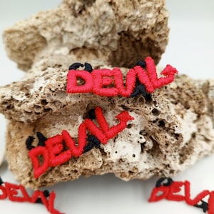 May include: Red embroidered patches with the word "DEVIL" in black outlined letters. The patches are on a rough, light brown rock surface.