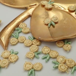 May include: A collection of small, yellow, embroidered flower embellishments with green stems. The flowers are scattered on a white surface, with a gold metal clover-shaped object in the background.