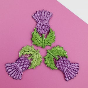May include: Three embroidered thistle patches in purple and green. The thistles are arranged in a triangular pattern.