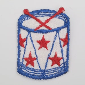 May include: A blue and white embroidered patch of a drum with red stars and drumsticks.
