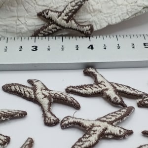 May include: A group of six small, brown embroidered bird appliques. The birds are about 1 inch long and have white stitching. A ruler is in the background for size reference.