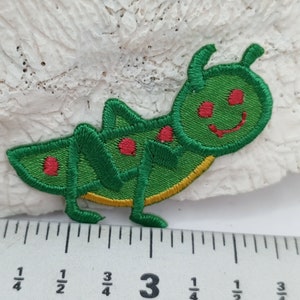 May include: Green embroidered iron-on patch of a cartoon caterpillar with red spots and a yellow outline. The caterpillar is approximately 7.5 cm long.