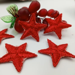 May include: A collection of red embroidered star patches with a white border, scattered on a white surface. A cluster of red glass grapes is in the background. The stars are a bright red colour and appear to be handmade.