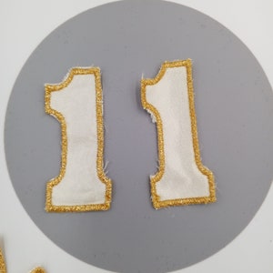 Könnte beinhalten: Zwei weiße Filzpatches mit goldener Stickerei, in Form der Zahl 11.