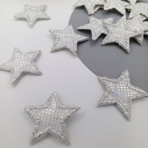 May include: A collection of silver glitter star-shaped fabric patches. The stars are scattered on a white surface with a few on a grey surface.