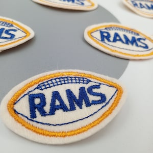 May include: A white felt football patch with a gold outline and the word "RAMS" embroidered in blue.