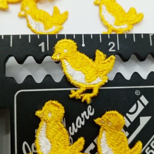 Pode incluir: Um conjunto de seis patches de feltro amarelo em forma de pintinhos para passar a ferro. Cada pintinho tem olhos brancos e um bico branco. Os patches estão dispostos numa superfície preta com uma régua.