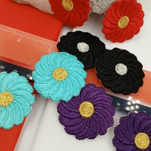 Puede incluir: Surtido de parches bordados de flores de colores en turquesa, morado, negro y rojo. Los parches tienen un diseño de espiral y un centro amarillo.