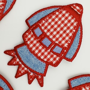 Könnte beinhalten: Roter und weißer Gingham-Stoff-Raketen-Patch mit blauem Jeansbesatz. Die Rakete hat eine spitze Nase und einen Schwanz mit Flammen.