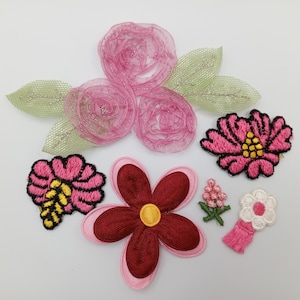 May include: Assortment of pink and red fabric flower appliques. The appliques are in various shapes and sizes, including a large pink rose, a smaller pink rose, a red flower with five petals, and a small pink flower with a yellow center.