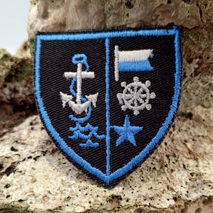 May include: A blue and black embroidered patch in the shape of a shield. The shield features a white anchor, a white steering wheel, a white star, and a blue and white flag.