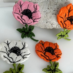 May include: Four embroidered fabric flower patches. One is pink, one is white, and two are orange. Each flower has a black center and green leaves.