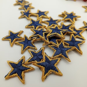 May include: A pile of navy blue fabric stars with gold trim. The stars are scattered on a white surface.