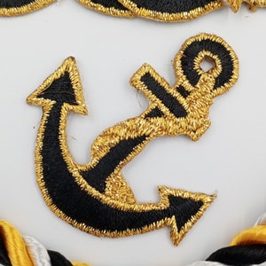 May include: A black and gold embroidered anchor patch. The anchor is outlined in gold thread and has a black center.