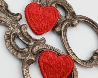 May include: Two ornate metal keychains with red felt hearts attached. The keychains are silver and have a detailed design.