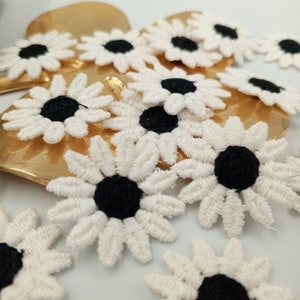 May include: A collection of white crocheted daisy flower appliques with black centers. The appliques are scattered on a gold surface.