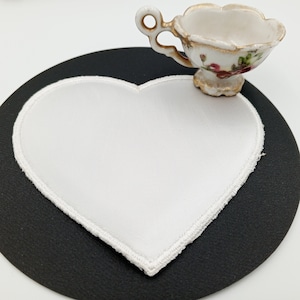 May include: A white heart-shaped coaster with a white stitched border sits on a black round surface. A small white teacup with a floral design sits on top of the coaster.