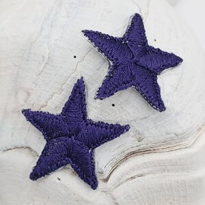 May include: Two purple embroidered star shapes on a white background.