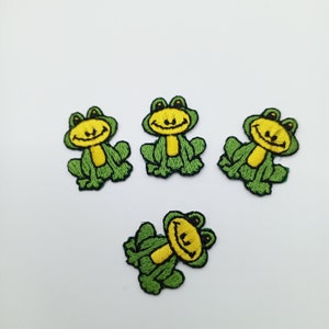 May include: Four green embroidered iron-on patches of smiling frogs. Each frog has yellow eyes, a yellow smile, and a yellow belly.