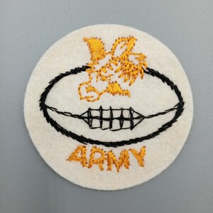 May include: A white felt patch with a black outline of a football and a gold embroidered image of a mascot. The word "ARMY" is embroidered in gold below the football.