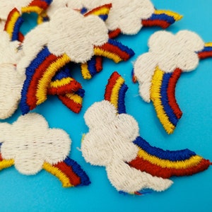 May include: A set of white felt patches with embroidered rainbow designs. Each patch features a white cloud with a rainbow arc underneath. The rainbows are embroidered in red, yellow, and blue thread.