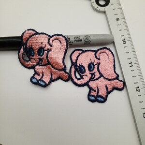 May include: Two pink embroidered iron-on patches of baby elephants with blue eyes and navy blue outlines.