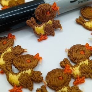 Pode incluir: Uma coleção de patinhos de feltro bordados em marrom e amarelo com bicos e pés laranja. Os patinhos estão dispostos em círculo sobre uma superfície branca.