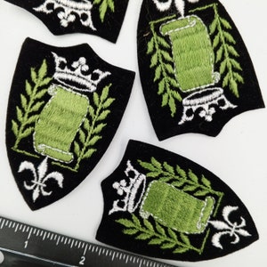 May include: Four black velvet patches with white and green embroidery. Each patch features a scroll with a crown on top, surrounded by green leaves. The scroll is blank.