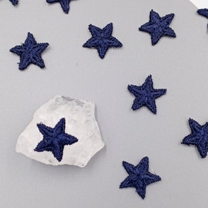 May include: A collection of navy blue embroidered star shapes on a light grey surface. The stars are scattered randomly and some are on a clear quartz crystal.