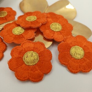 Könnte beinhalten: Eine Gruppe von sechs orangen Filzblumen-Patches mit goldenen Zentren. Die Patches sind in einer Gruppe auf einem goldenen Hintergrund angeordnet.
