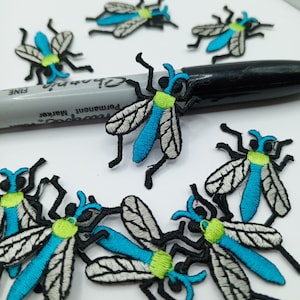 Pode incluir: Um conjunto de oito patches bordados de ferro azul e preto com um design de mosca estilizado. As moscas têm um corpo azul, pernas pretas e asas brancas com veias pretas.