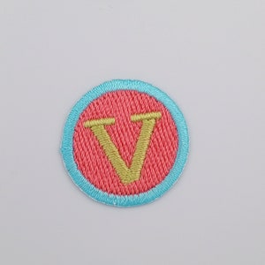 May include: An embroidered patch with a red centre and a blue border, featuring a gold letter V.