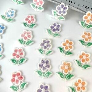 May include: Assortment of colorful embroidered flower appliques with white outlines. The flowers are in shades of pink, blue, purple, orange, and yellow. Each flower has five petals and is surrounded by two green leaves.