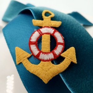May include: A yellow embroidered anchor with a red and white life preserver around the top. The anchor is on a teal blue velvet background.