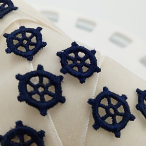 May include: A close-up of a white ribbon with navy blue embroidered nautical wheel designs. The ribbon is perfect for adding a touch of nautical charm to crafts, gifts, and home decor.